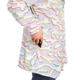 A close-up of a person wearing a light lavender long-sleeve tunic top with a pastel multicolor wavy abstract pattern. The person is shown from the side, with a hand reaching into a side pocket, with dark blue jeans visible at the bottom.