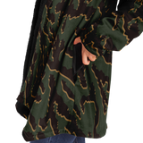 A close-up of a person wearing a long camouflage-patterned jacket with a black fleece lining and side pockets, with a hand reaching into one of the pockets.